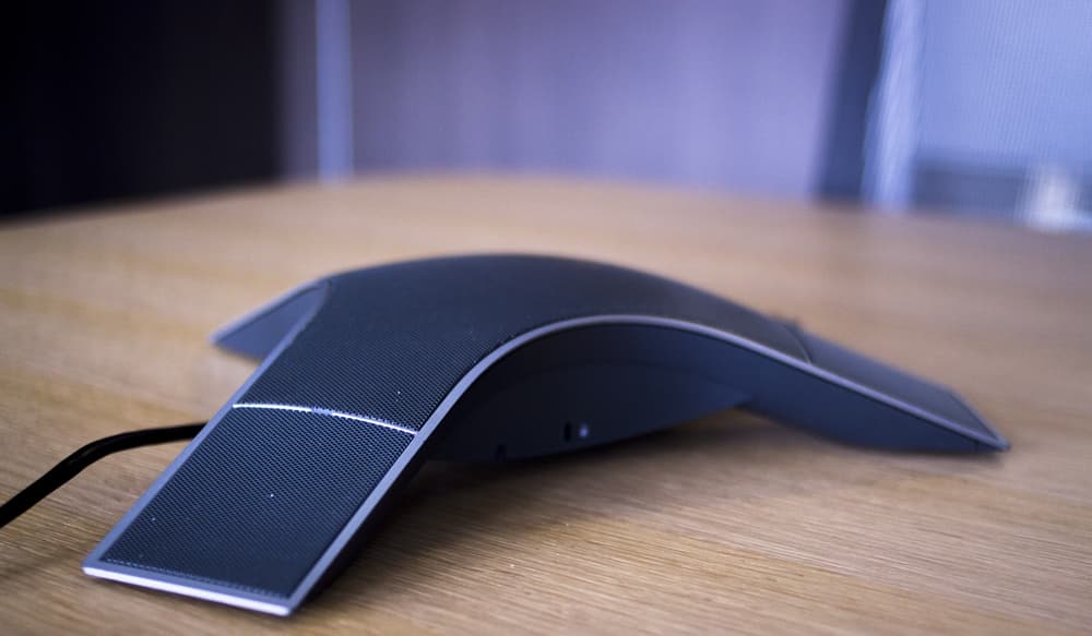 A black conference speakerphone with a sleek, curved design sits on a wooden table, with a cable connected to it.