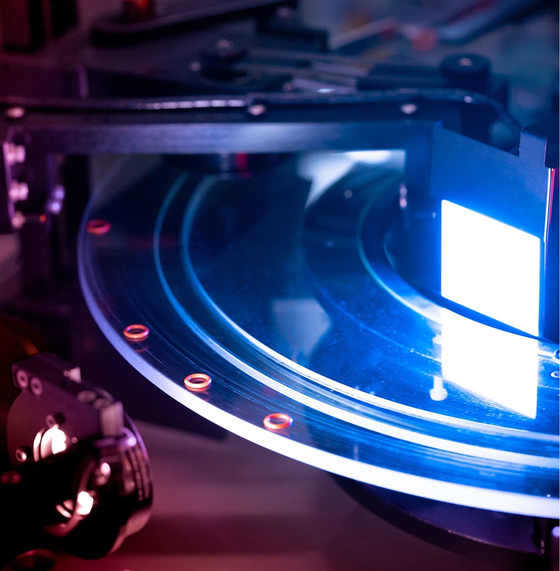 Detailed close-up of a semiconductor wafer illuminated by bright light inside a testing machine, highlighting intricate surface details and advanced technology used for quality inspection in electronics manufacturing.