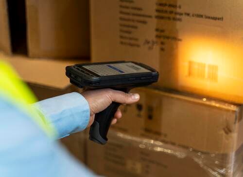 A worker uses a handheld device to scan a barcode on a cardboard box in a warehouse, reflecting strict manufacturing process controls and adherence to ESD-safe protocols during the assembly of electronic and electromechanical components.